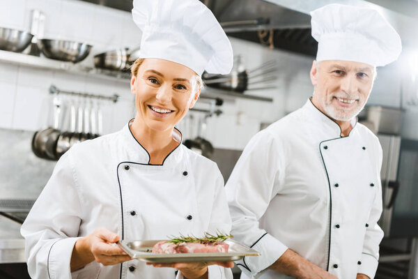 female and male chefs in uniform with meat steak on plate looking at camera in restaurant kitchen