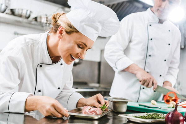 smiling female chef holding meat dish while colleague in uniform cooking on background in restaurant kitchen