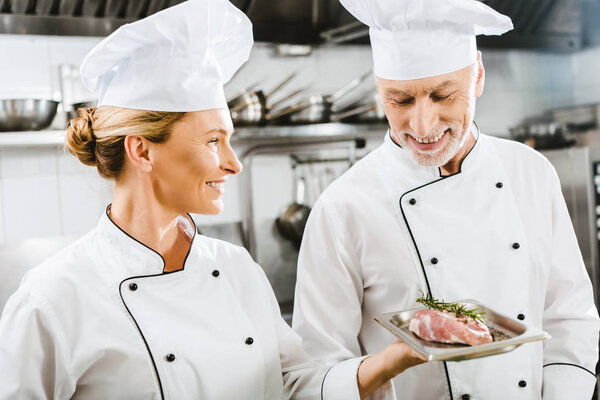 smiling female chef in uniform presenting meat dish to colleague in restaurant kitchen