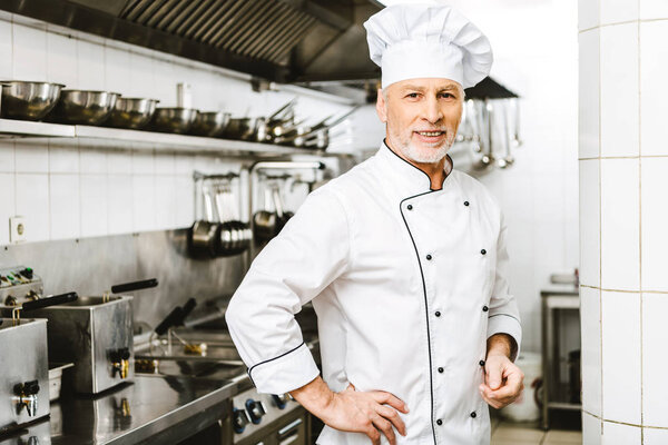 handsome smiling male chef in uniform and cap looking at camera in restaurant kitchen