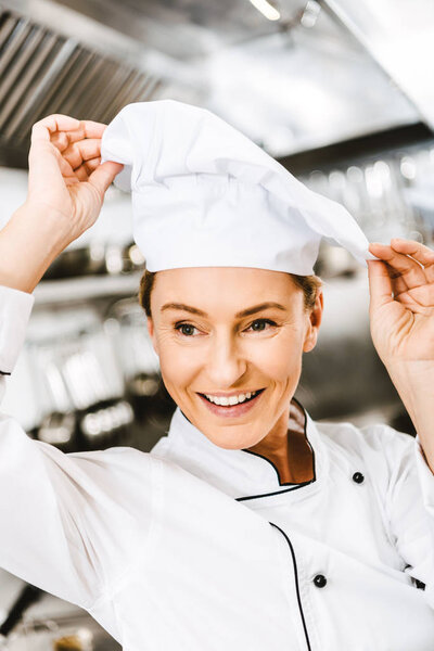 beautiful smiling female chef adjusting cap in restaurant kitchen