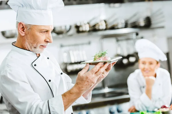 沉思的男厨师在制服举行肉菜在盘子与同事在后台在餐厅厨房 — 图库照片