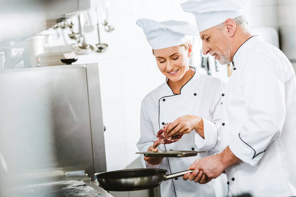 female and male chefs in uniform cooking meat on pan in restaurant kitchen with copy space