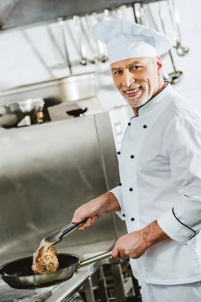 笑顔の男性シェフがレストランのキッチンで肉ステーキを焙煎しながらカメラ目線 — ストック写真