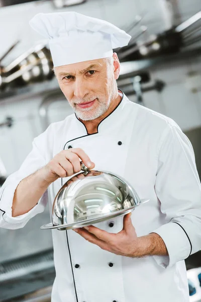 英俊的男性厨师在制服看相机 拿着服务托盘圆顶在餐厅厨房 — 图库照片