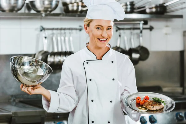 レストランのキッチンで料理に肉のトレイを提供からドームを保持している制服を着た美しい笑みを浮かべて女性シェフ — ストック写真