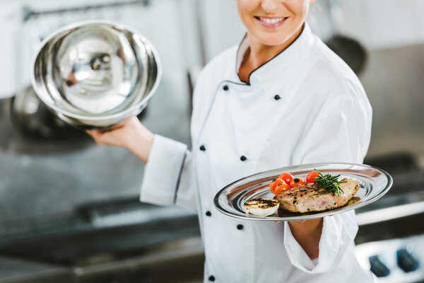 cropped view of female chef in uniform holding dome from serving tray with meat dish in restaurant kitchen
