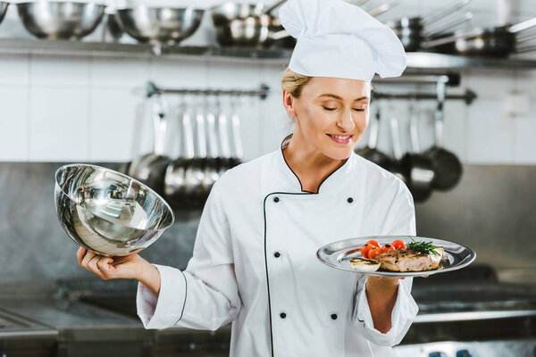 selective focus of beautiful female chef in uniform holding serving tray with meat dish in restaurant kitchen