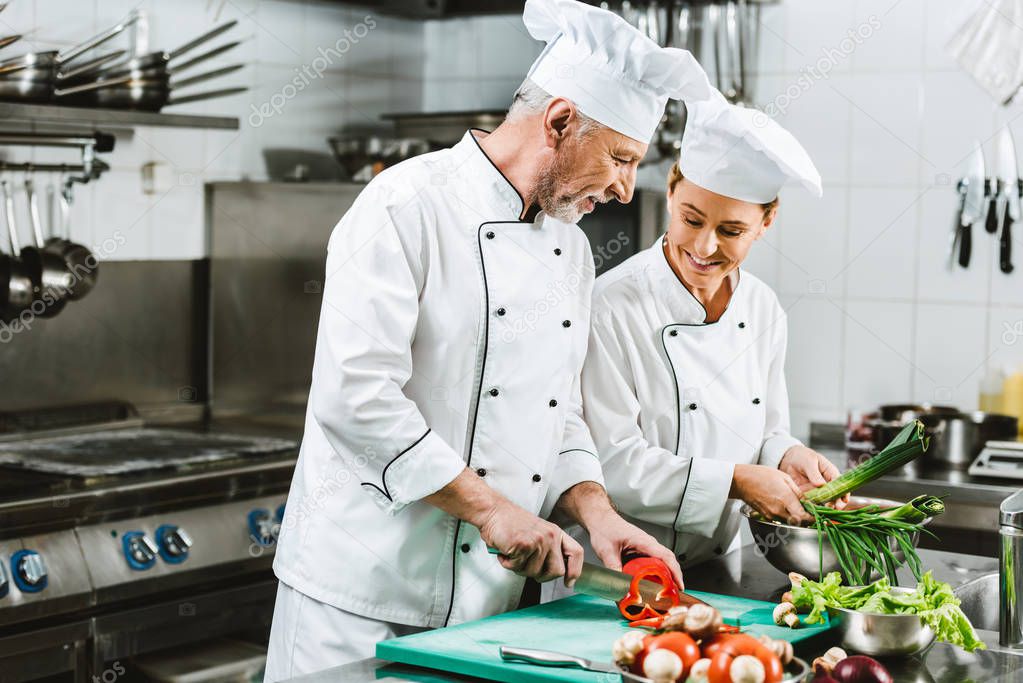 smiling female and male chefs in uniform and hats cooking in restaurant kitchen