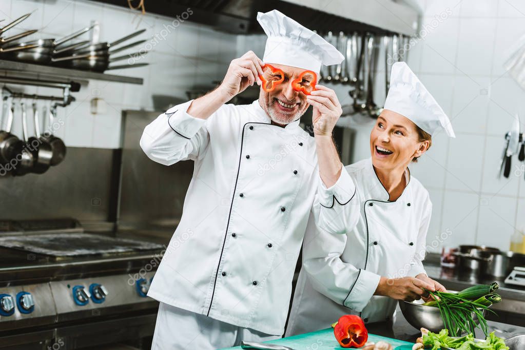 smiling female cook looking at male chef holding pepper slices in front of face in restaurant kitchen