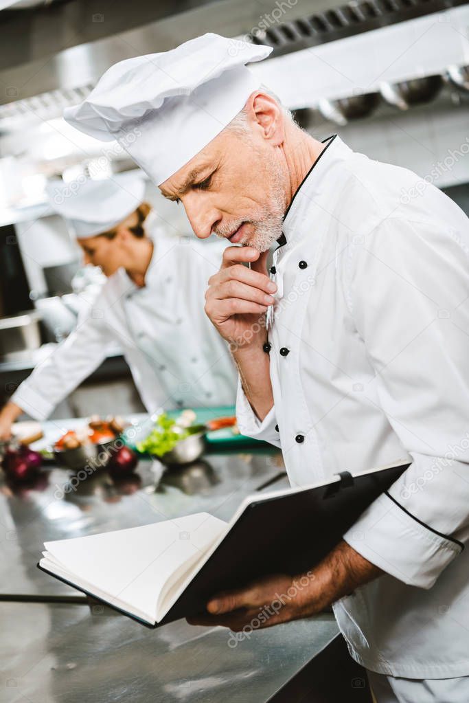 handsome thoughtful male chef propping chin with hand while reading recipe book in restaurant kitchen