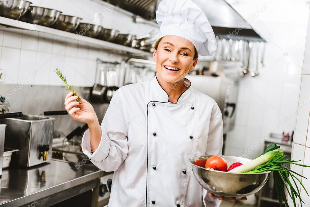 beautiful smiling female chef in uniform holding rosemary and bowl with vegetables in restaurant kitchen