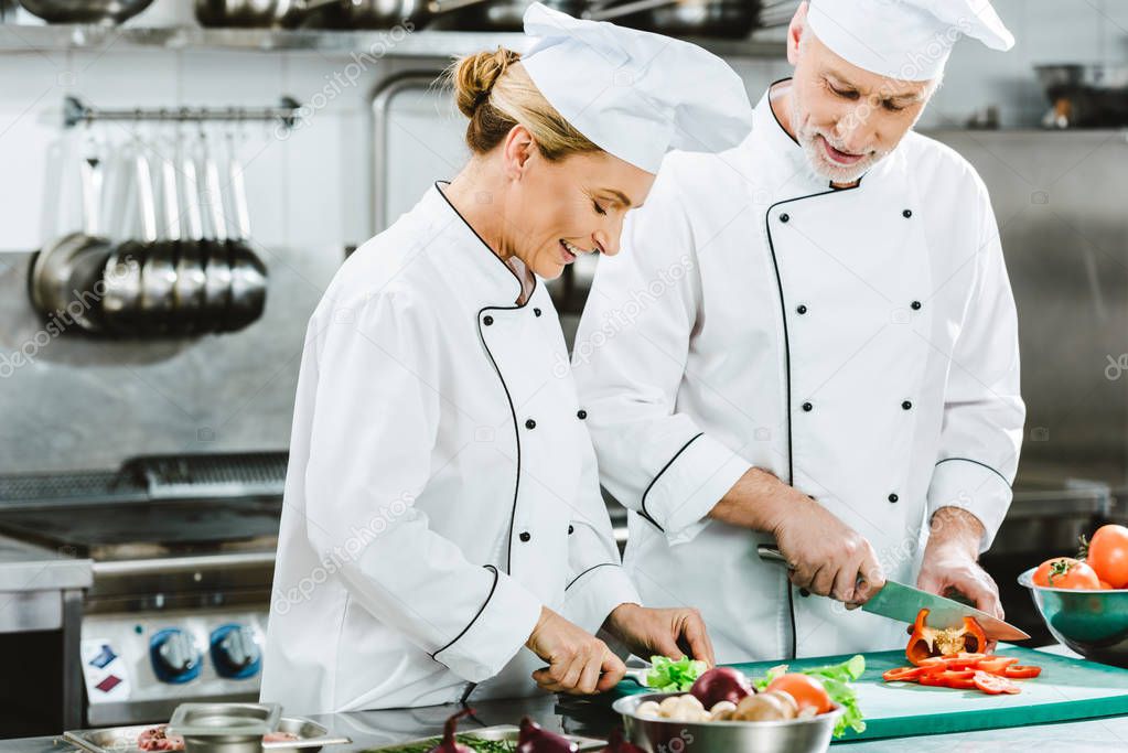 smiling female and male chefs in uniform cutting ingredients while cooking in restaurant kitchen