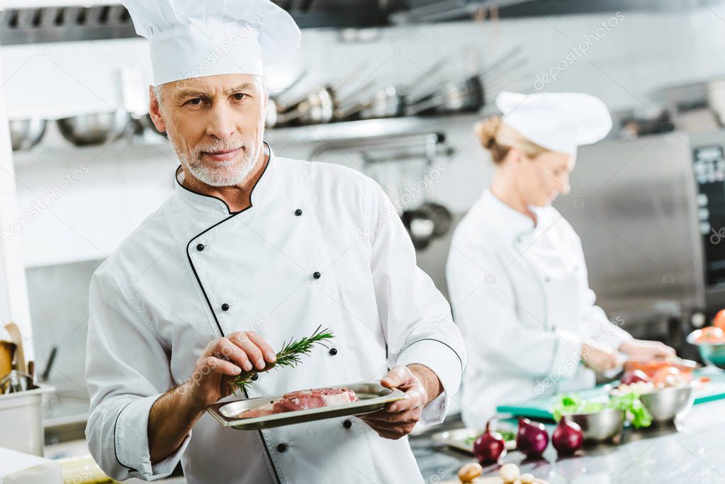 male chef in uniform looking at camera and holding meat dish on plate with colleague cooking on background in restaurant kitchen