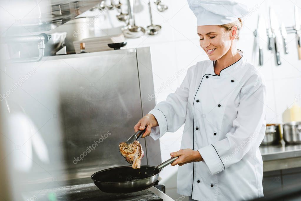 beautiful smiling female chef in uniform cooking meat in restaurant kitchen