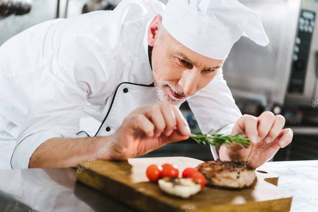 selective focus of male chef in uniform decorating meat steak with rosemary in restaurant kitchen