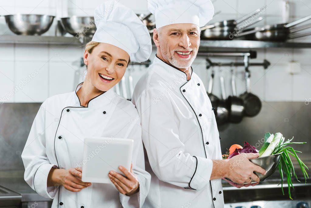 female and male chefs in iniforms looking at camera and using digital tablet while cooking in restaurant kitchen