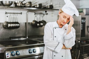 beautiful tired female chef in uniform having headache in restaurant kitchen with copy space clipart