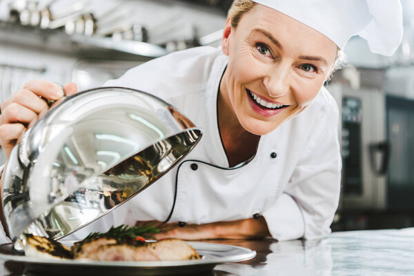 beautiful female chef in uniform looking at camera and holding dome from serving tray with meat dish in restaurant kitchen