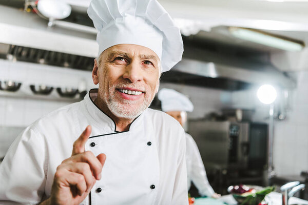 handsome smiling male chef in uniform and cap doing idea gesture in restaurant kitchen