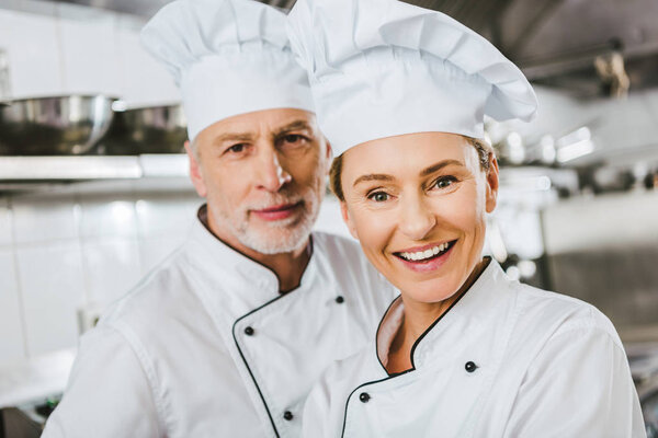 female and male chefs in uniforms looking at camera and smiling at restaurant kitchen