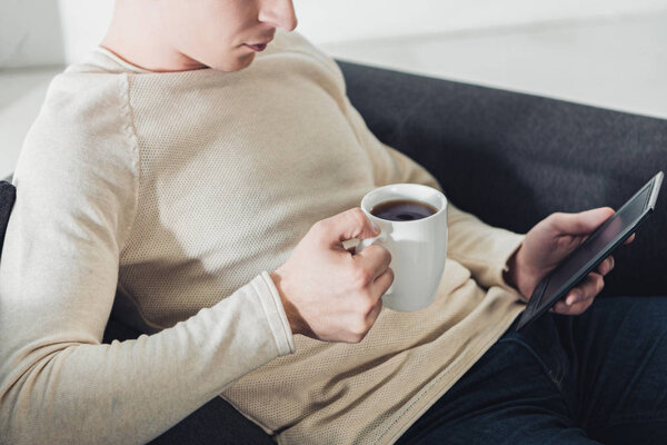 cropped view of man holding cup with coffee and reading ebook 