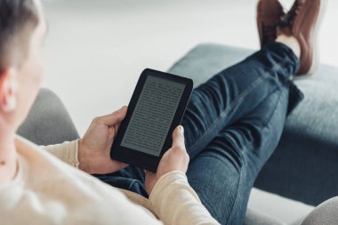 cropped view of man reading e-book while sitting in armchair at home clipart