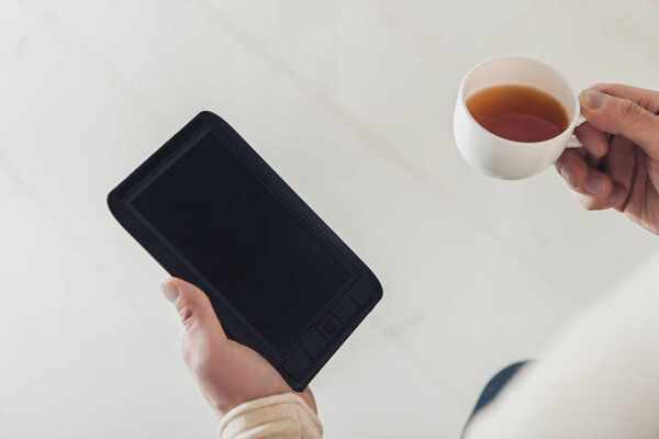 cropped view of ebook with blank screen and cup of tea in hands of man at home