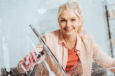Wonderful senior woman using glass wiper while cleaning window clipart