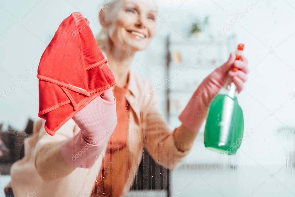 Joyful senior woman cleaning window with spray and red rag