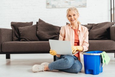 Evi temizlerken dizüstü kullanarak lastik eldiven içinde üst düzey kadın gülüyor