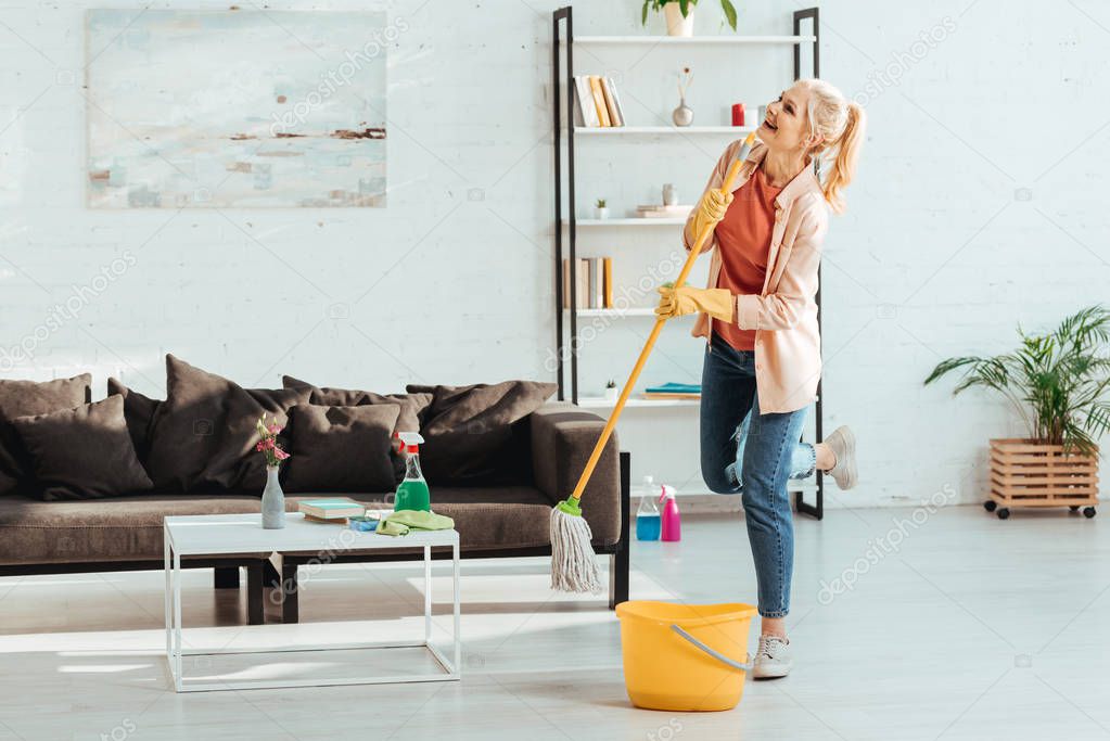 Laughing senior woman posing on one leg while cleaning floor with mop
