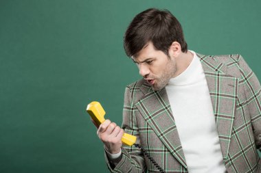 handsome man in vintage clothes shouting at retro telephone isolated on green with copy space clipart