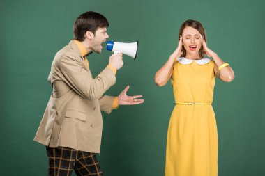 woman in vintage clothes covering ears with hands while angry man yelling in loudspeaker isolated on green clipart
