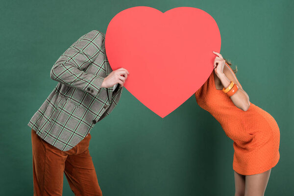 man and woman covering faces with heart shaped paper card isolated on green