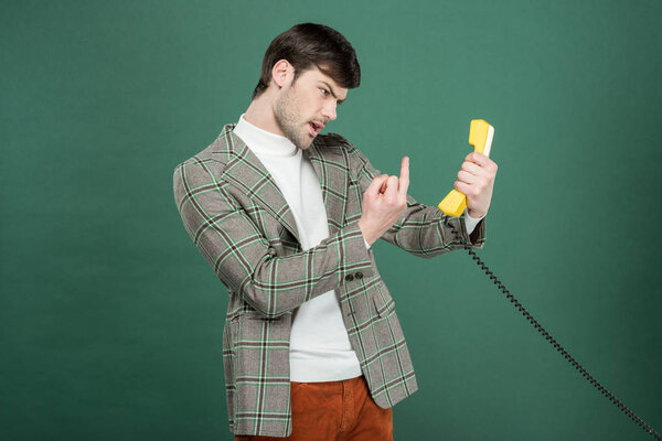 angry handsome man in vintage clothes showing middle finger in front of retro telephone isolated on green