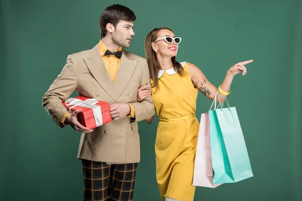 man in vintage clothes with gift box and woman pointing with finger while holding shopping bags isolated on green