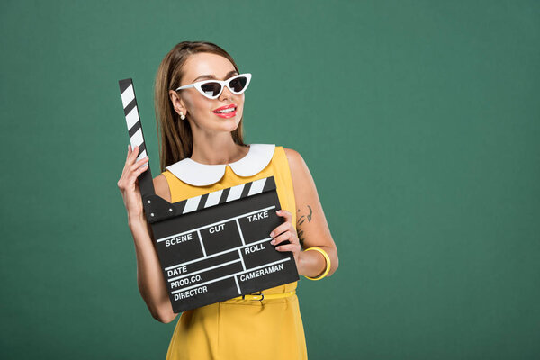 beautiful smiling stylish woman in yellow dress and sunglasses holding film clapperboard isolated on green