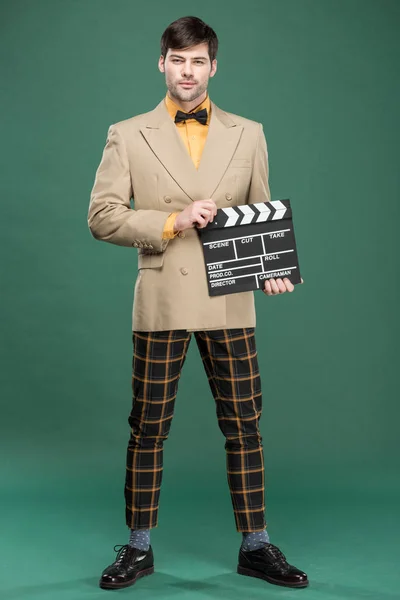 handsome man in vintage clothes looking at camera and holding film clapperboard on green background