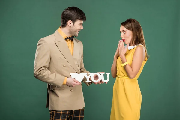 man presenting decorative 'i love you' sign to beautiful surprised woman in vintage clothes isolated on green