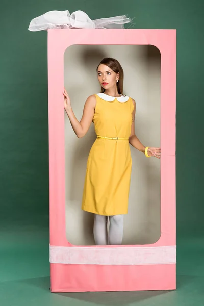 緑の背景に弓で装飾的なピンクの箱でポーズをとって黄色のドレスでスタイリッシュな美人 — ストック写真