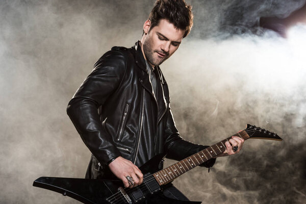 handsome rocker in leather jacket playing electric guitar on smoky background