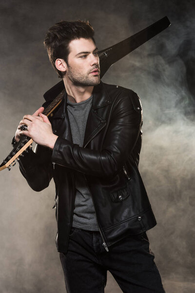 handsome rocker holding electric guitar and looking away on smoky background