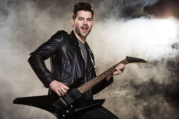 handsome rocker in leather jacket playing electric guitar on smoky background