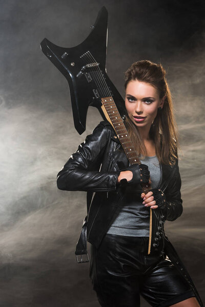 beautiful woman in leather jacket looking at camera and posing with electric guitar on smoky background