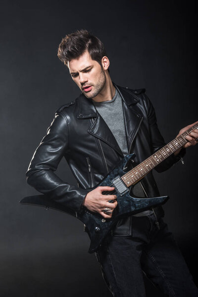 handsome rocker in leather jacket playing electric guitar isolated on black