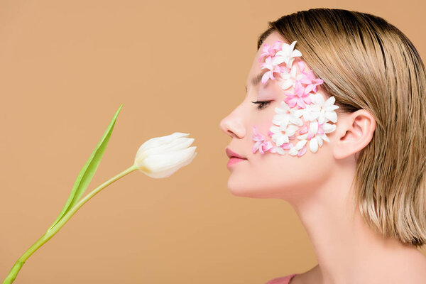 side view of elegant woman with flowers on face smelling tulip isolated on beige