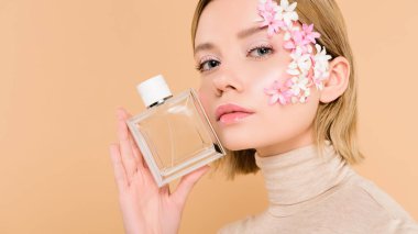 confident woman with flowers on face holding bottle of perfume isolated on beige clipart