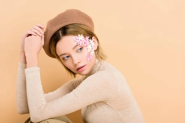 ベージュに分離されたベレー帽を身に着けている顔に花を持つ魅力的な女性 — ストック写真
