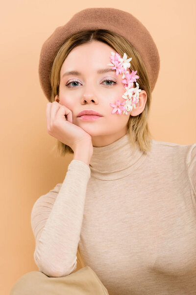 serious woman with flowers on face looking at camera isolated on beige 